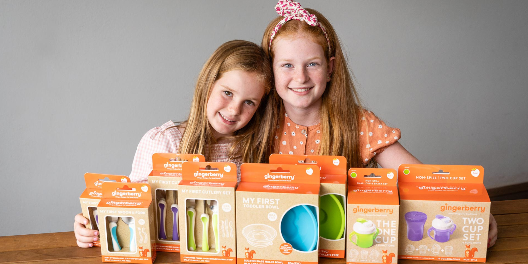 two girls displaying the gingerberry product range including training cutlery , bowls ,weaning spoons and sippy cups.