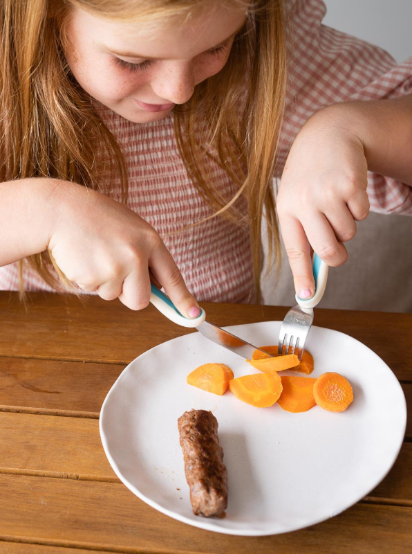   A todldler using the  My First Cutlery' set , including a knife, fork, and spoon. Each utensil features ergonomic handles with finger indents for ease of use, helping young children develop self-feeding skills. The stainless-steel fork has rounded prongs, the spoon has a shallow bowl, and the knife is designed to be safe for small hands, all perfectly sized for little mouths. The soft, overmoulded handle materials provide added grip and comfort.